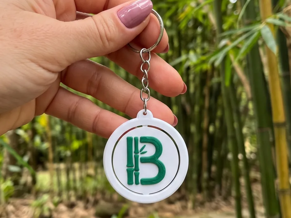 Chaveiro NFC redondo, branco com a letra B verde em forma de bambu, segurado na mão diante de um bambuzal