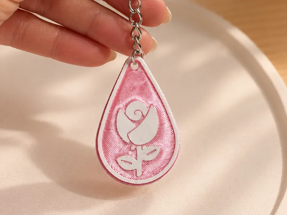Chaveiro NFC em gota, rosa e branco, com uma rosa em relevo, preso na mão