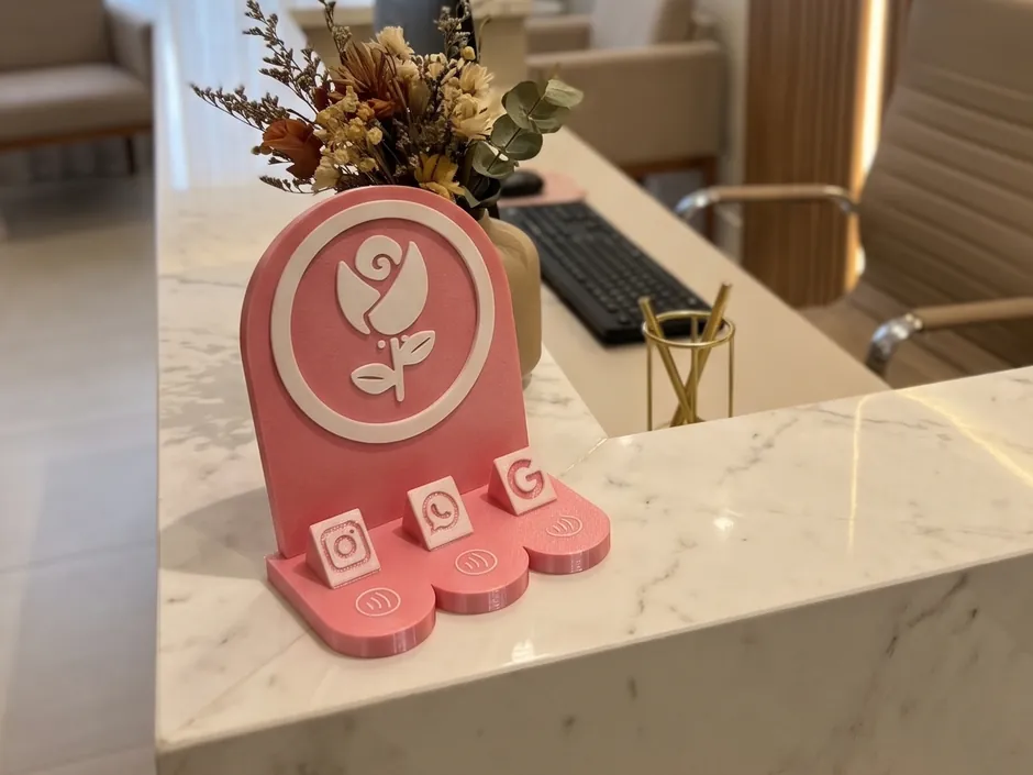 Totem de balcão rosa e branco com uma rosa em relevo e três botões, Instagram, WhatsApp e Google, cada um com o sinal de aproximar o celular, sobre o balcão de mármore da recepção de uma clínica